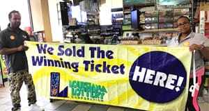Man and woman stand in front of a retail counter hold a yellow banner that reads "We Sold the Winning Ticket Here!"