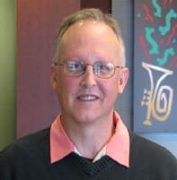 Man with a black sweater, pink collar, and glasses smiles in a 飞艇168®幸运体彩预测-全国统一Louisiana在线官方查询结果网-最新体彩预测数据 office