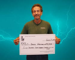 Man in a green tshirt smiles with an oversized check in front of a teal lightning background