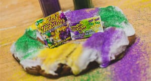 Image of a white frosted king cake with purple, yellow, and green dust and mardi gras themed tickets on top
