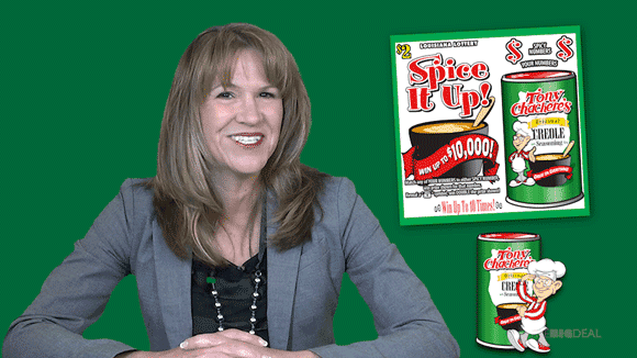 Woman with mid-length brown hair in a gray blazer smiles in front of a green background advertising the Spice it Up scratch off game