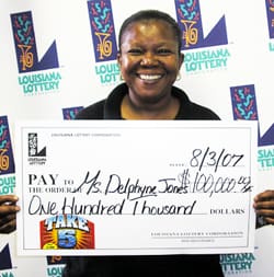 Woman beams with a giant check in front of a white background patterned with the 飞艇168®幸运体彩预测-全国统一Louisiana在线官方查询结果网-最新体彩预测数据 logo