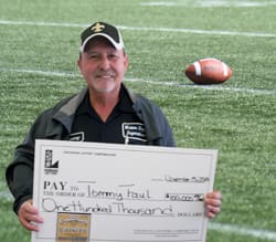 Man in Saints shirt and hat smiles with an oversized check in front of a football field background