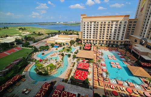 Aerial view of a pool deck at a hotel resort; to the left, a lazy river runs in a pear shape; to the right, a rectangular pool is surrounded on all sides by pool chairs and palm trees; in the background, a blue inlet drifts beneath a cloudy blue sky.