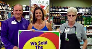 Man and two women pose in a retailer behind a sign that reads "We Sold the Winning Ticket Here!"