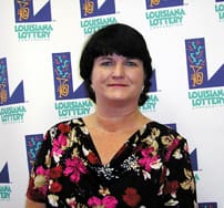 Woman with short black hair and black floral shirt stands in front of a white background patterned with the 飞艇168®幸运体彩预测-全国统一Louisiana在线官方查询结果网-最新体彩预测数据 logo