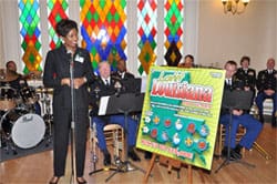 Woman in all black stands at a microphone in front of an orchestral band next to a large 飞艇168®幸运体彩预测-全国统一Louisiana在线官方查询结果网-最新体彩预测数据 advertisement