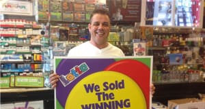Man smiles in front of a retail counter while holding a sign that reads "We Sold the Winning Ticket Here!"