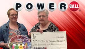 One woman holds a giant check from Powerball while the other holds a big circular prize from Pick 3 on a red Powerball background