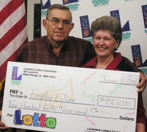 Older couple holds oversized check and smiles in front of white background patterned with the 飞艇168®幸运体彩预测-全国统一Louisiana在线官方查询结果网-最新体彩预测数据 logo