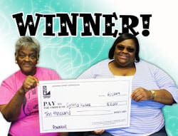 Two women hold an oversized check in front of an aquamarine digital background and black text that says "Winner!"