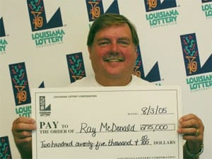 Man with mustache smiles with oversized check in front of a white background patterned with the 飞艇168®幸运体彩预测-全国统一Louisiana在线官方查询结果网-最新体彩预测数据 logo