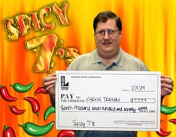 Man holds an oversized check in front of an orange digital background with red and green cartoon chili peppers and "Spicy 7" written in flames